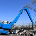 Blue material handler sorting scrap metal at a recycling yard